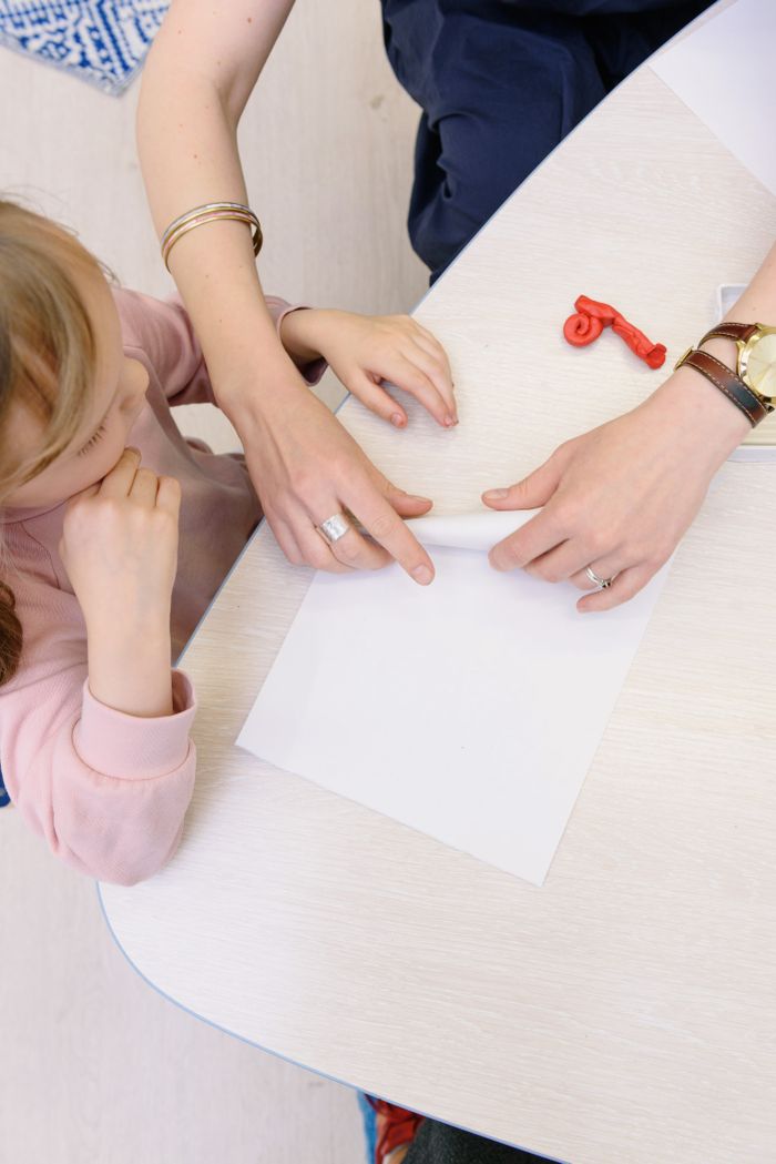 Top view of an adult guiding a child in a crafts activity at a table indoors.